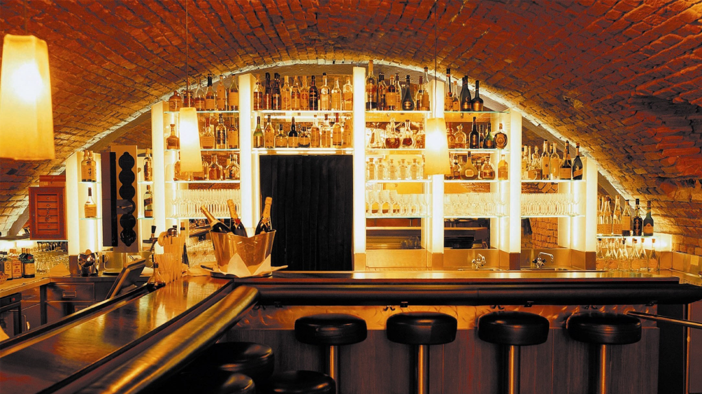 Ein Bild der gemütlichen Bar des Tigerpalasts: Im Vordergrund stehen Barhocker und eine Theke, im Hintergrund elegante Regale mit verschiedenen Flaschen und Gläsern.