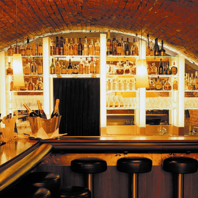 Ein Bild der gemütlichen Bar des Tigerpalasts: Im Vordergrund stehen Barhocker und eine Theke, im Hintergrund elegante Regale mit verschiedenen Flaschen und Gläsern.