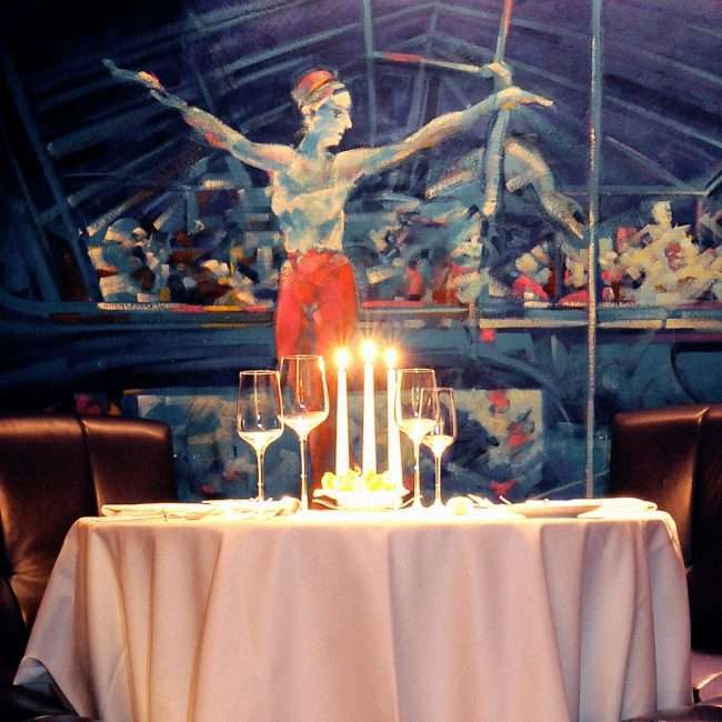 Zu sehen ist ein weiß gedeckter Tisch im Tigerpalast-Restaurant. An der Wand dahinter hängt ein malerisches Bild einer Akrobatin.